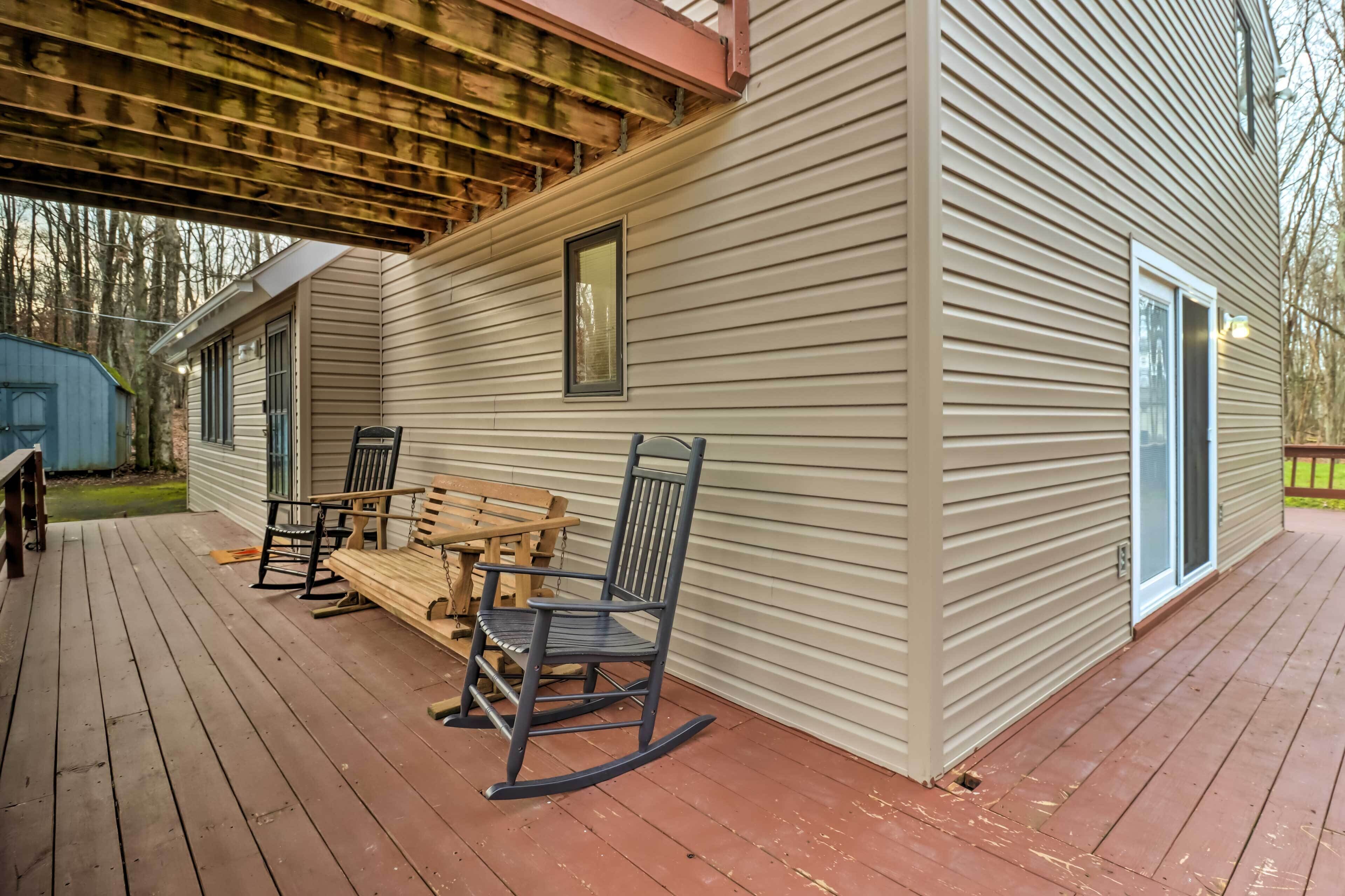 A covered deck with a wooden table and chairs, a rocking chair, and a small shed in the background.