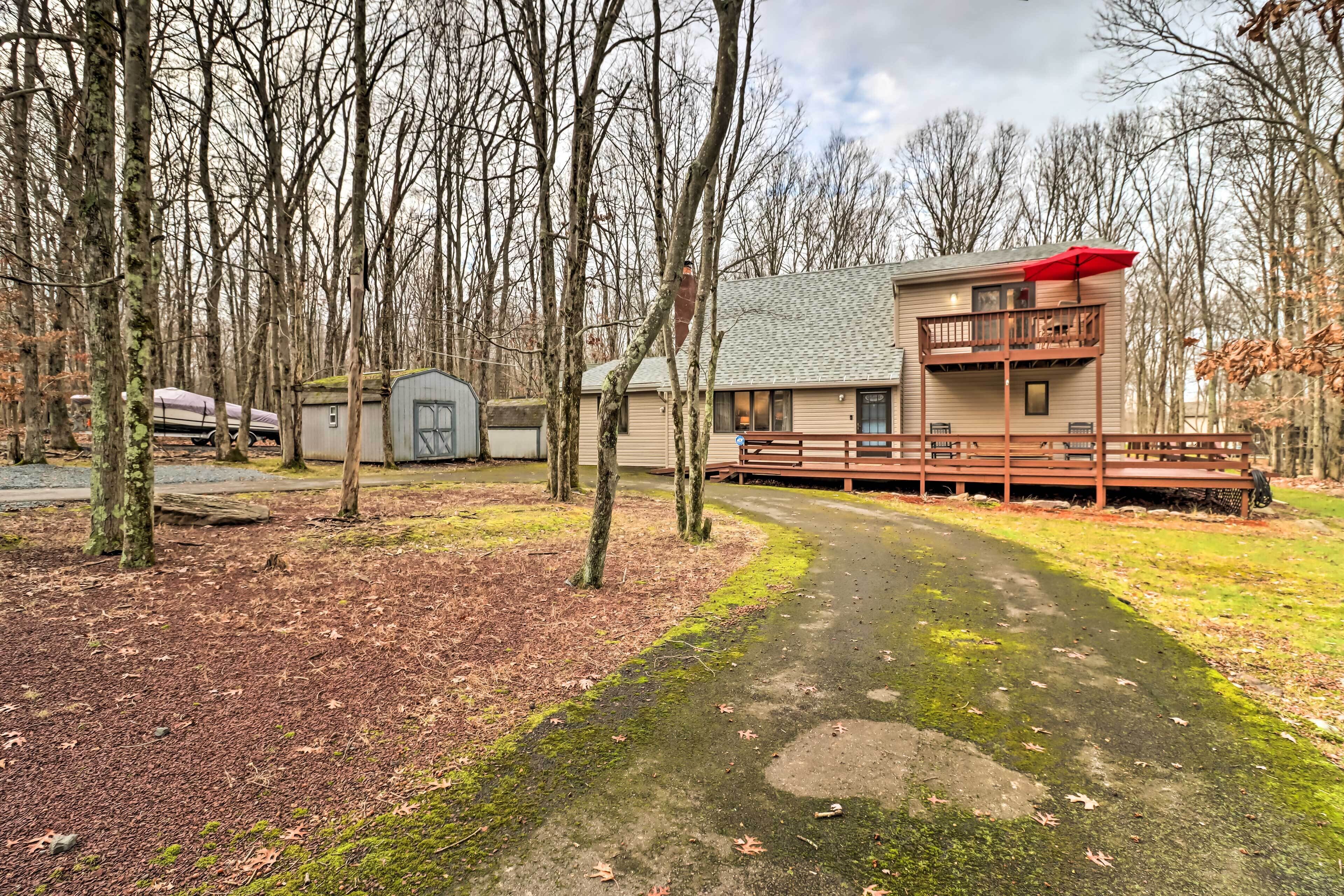 A cabin with a deck and a covered porch, surrounded by trees and a gravel path.
