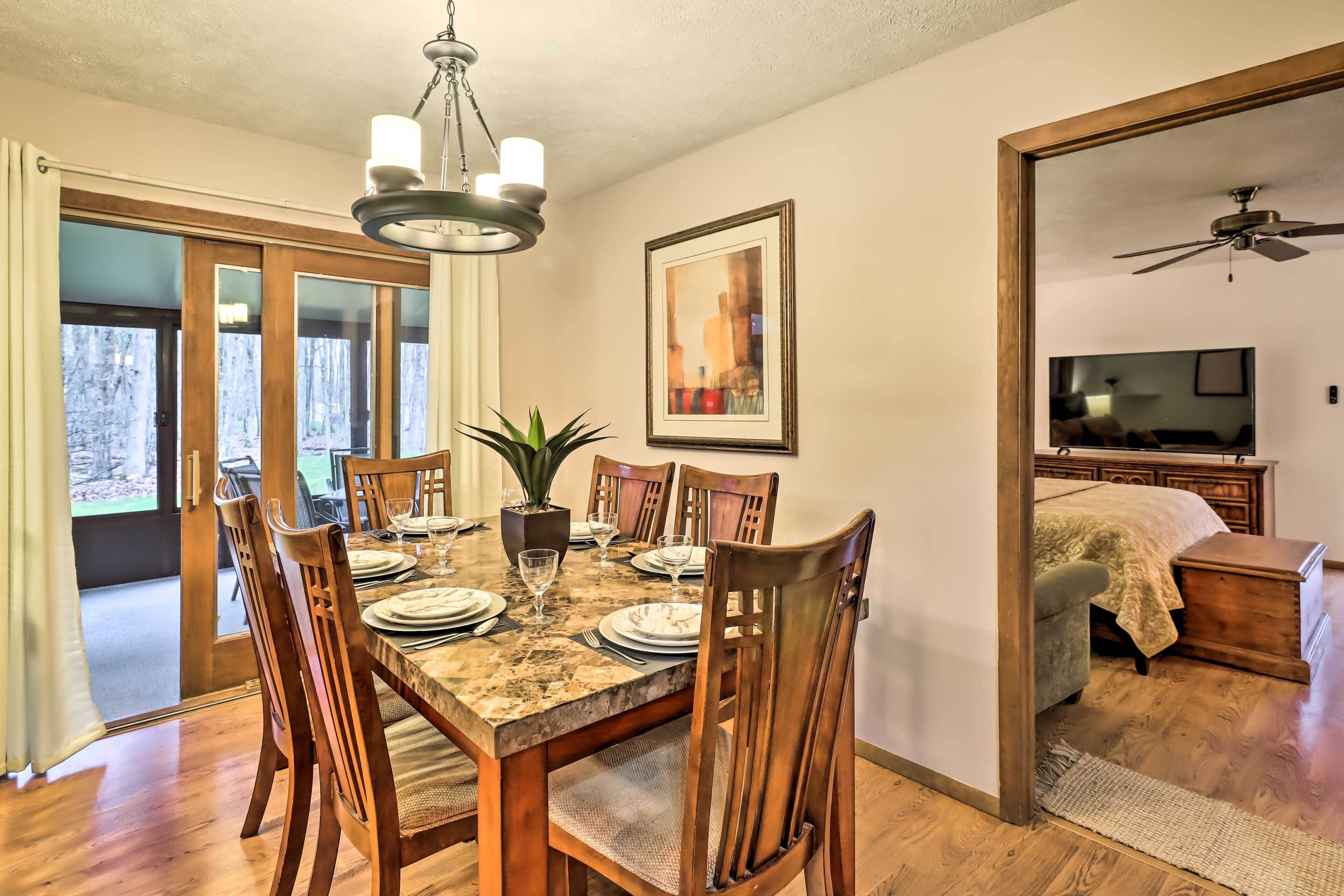 A dining area with a wooden table set for six, surrounded by chairs, a chandelier, and a potted plant.