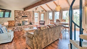 A spacious living room with a stone fireplace, wooden beams, and large windows offering a view of the outdoors. - Hot Tub, Game Room: Spacious Angel Fire Retreat! (Angel Fire)
