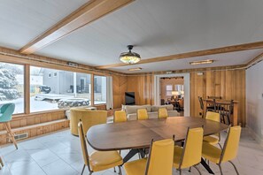 Een ruime eetruimte met een grote houten tafel en gele stoelen, een televisie en uitzicht naar buiten.