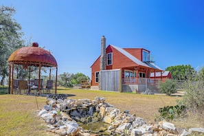 A red house with a stone chimney, a gazebo, and a small pond with rocks. - Pet-friendly Fredericksburg Cabin w/ Hot Tub! (Fredericksburg)