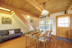 Ein gemütliches Interieur mit Holzbalkendecke, einem Esstisch mit Stühlen, einer Couch mit Kissen, einer Wanduhr und einer Tür mit Fenster.