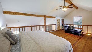 A bedroom with a sloped ceiling, wooden beams, a bed with a quilt, a leather sofa, and a window with blinds.