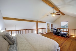 Ein Schlafzimmer mit schrägen Wänden, Holzbalken, einem Bett mit Patchworkdecke, einem Ledersofa und einem Fenster mit Jalousien.