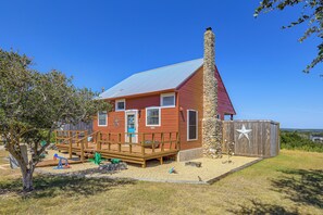 A red wooden house with a stone chimney, a wooden deck, and a fenced yard. - Pet-friendly Fredericksburg Cabin w/ Hot Tub! (Fredericksburg)