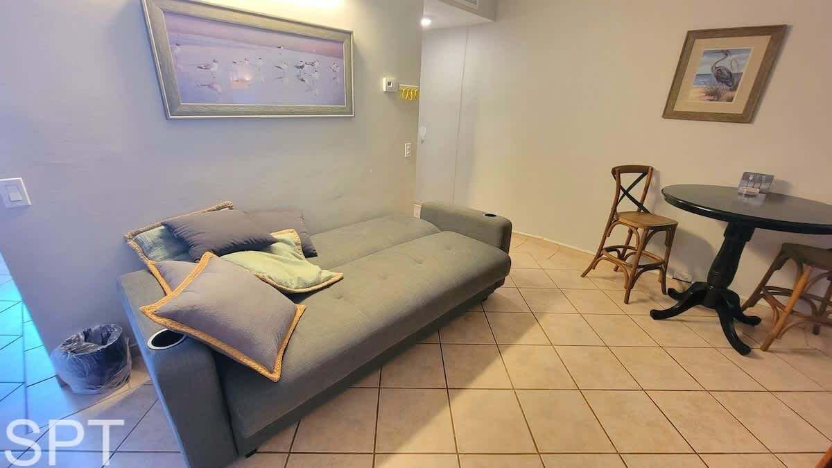 Una habitación con un sofá gris, dos sillas de madera, una mesita redonda y un cuadro en la pared.