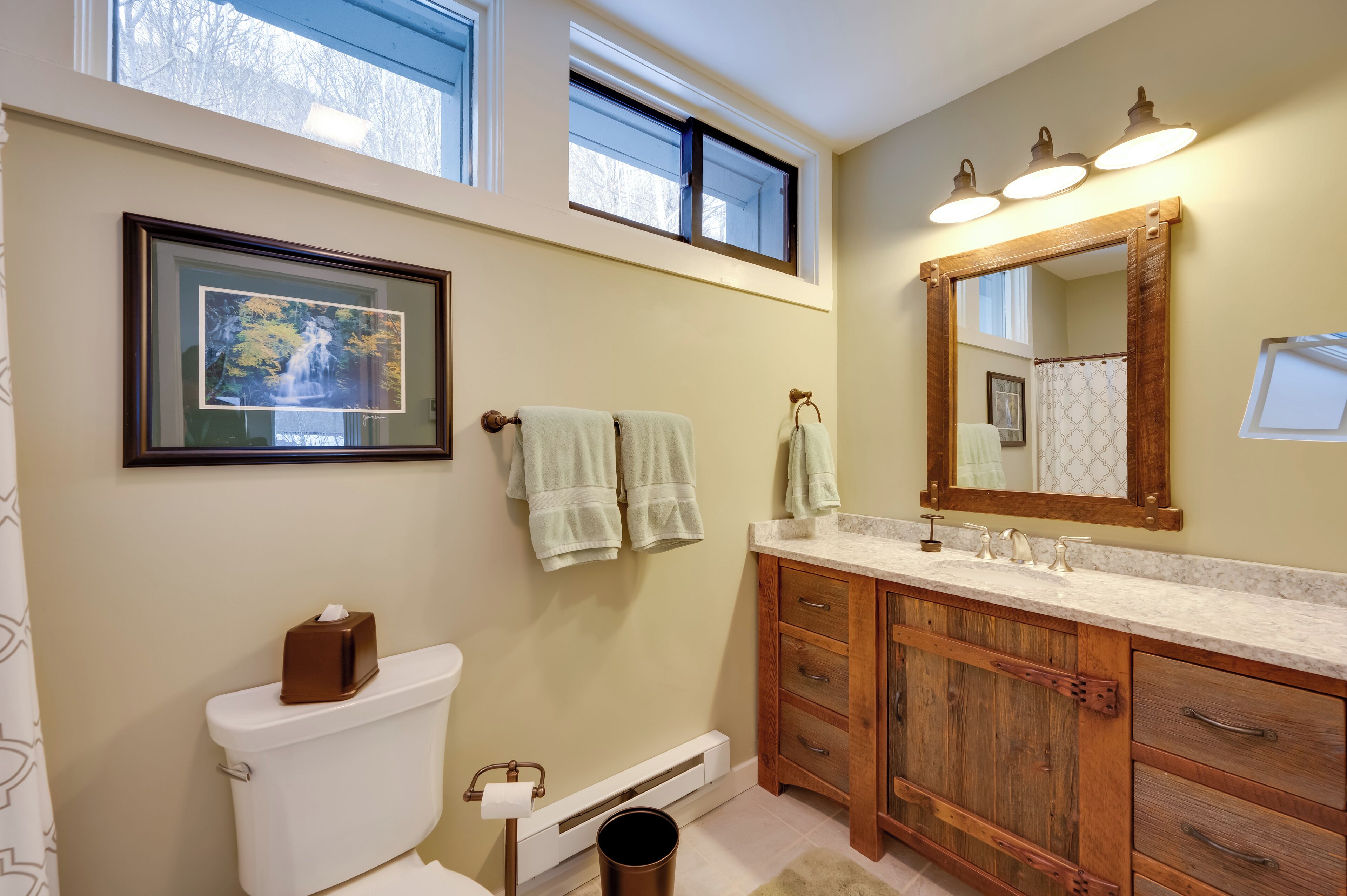 Une salle de bain comprenant des toilettes, un tableau encadré, un lavabo avec un miroir et une fenêtre située au-dessus du lavabo.