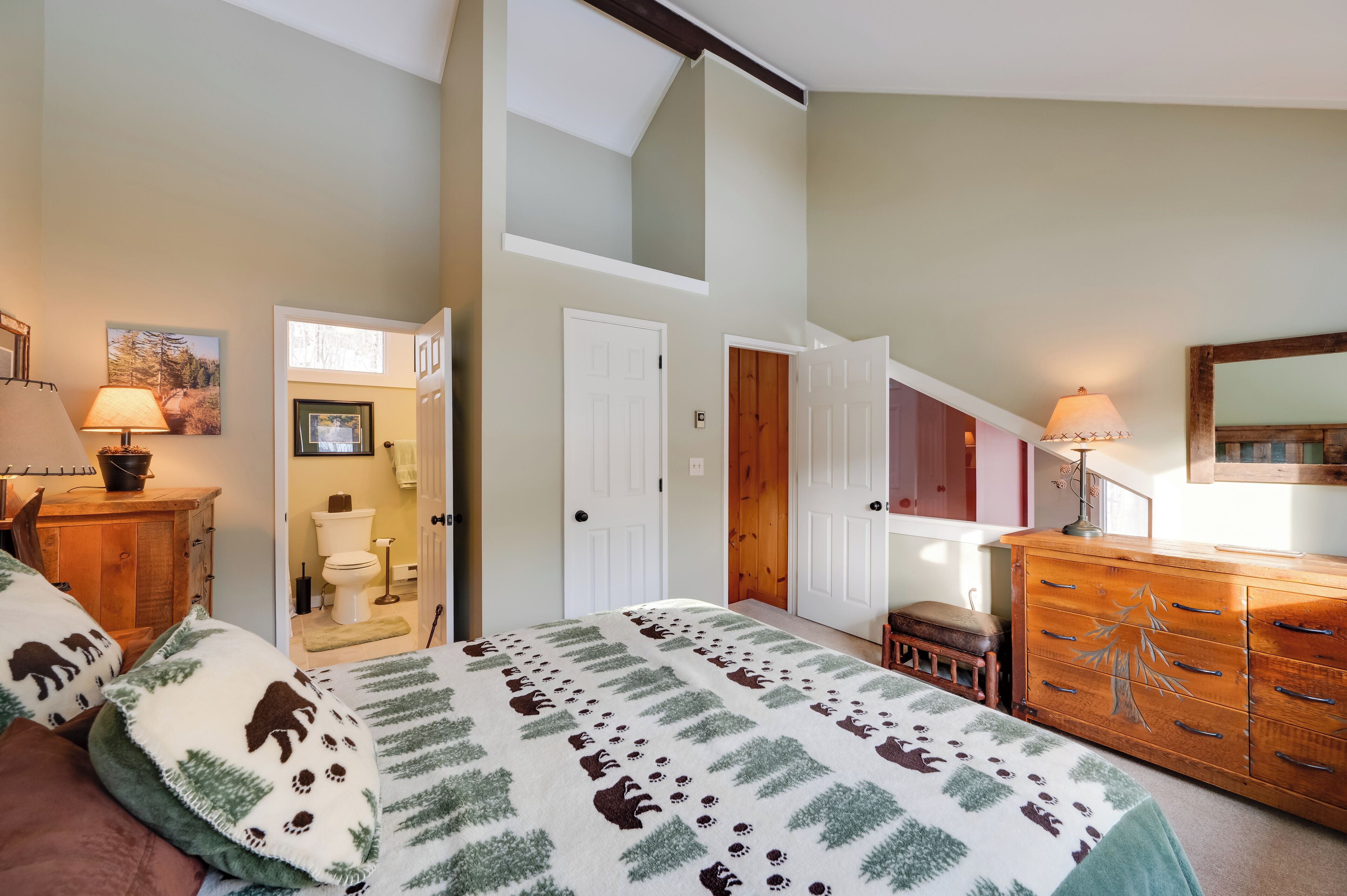 Une chambre à coucher comprenant un lit, une commode, une table de chevet, une lampe, une fenêtre et une porte donnant sur une salle de bain.