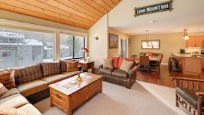 A spacious living area with a wooden ceiling, a large window, a sofa, a coffee table, and a kitchen area with a sign reading "LOON MOUNTAIN".