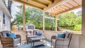 A covered patio with wicker furniture, a glass table, and a view of a house and hammock in the background. - Pet-friendly Pensacola Home w/ Sunny Backyard! (Pensacola)