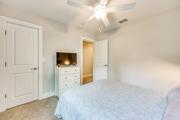 Une chambre à coucher avec un lit, un ventilateur de plafond, un téléviseur posé sur une commode et une porte donnant sur une autre pièce.