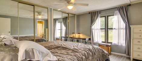 A bedroom with a large bed, a desk, and a chair, a ceiling fan, and a large window with curtains.