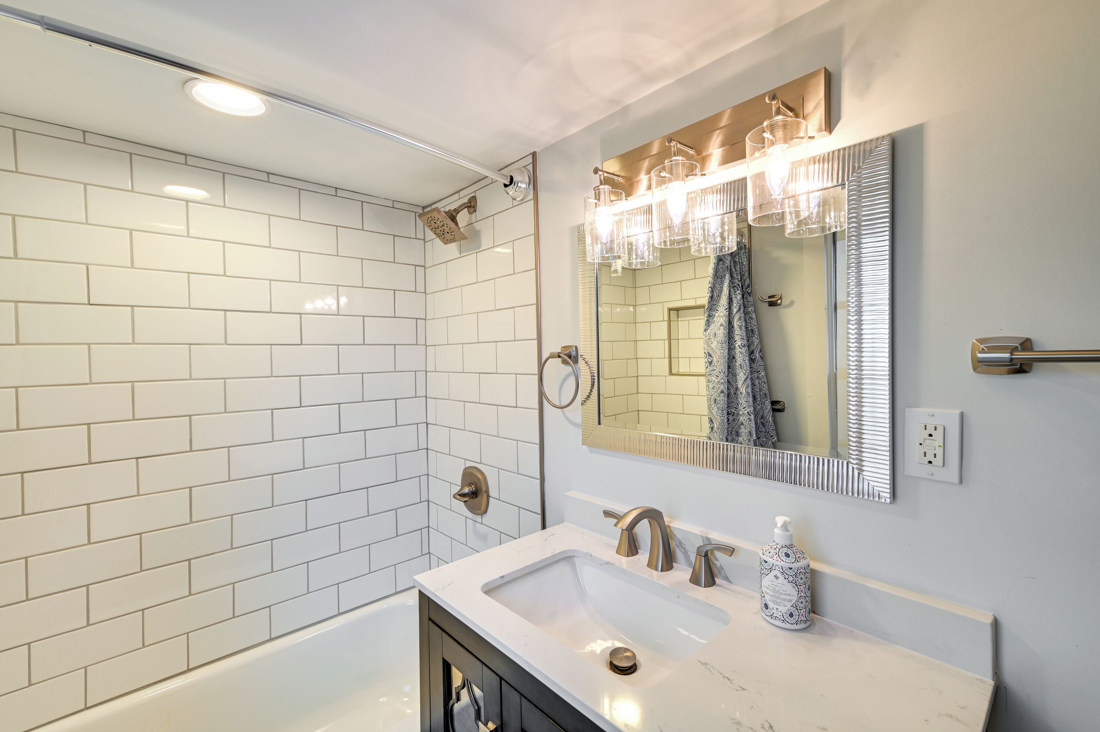 Ein modernes Badezimmer mit weißen, rechteckigen Fliesen, einem großen Spiegel mit Beleuchtung, einer Badewanne und einem Waschtisch mit Spüle und Armatur.