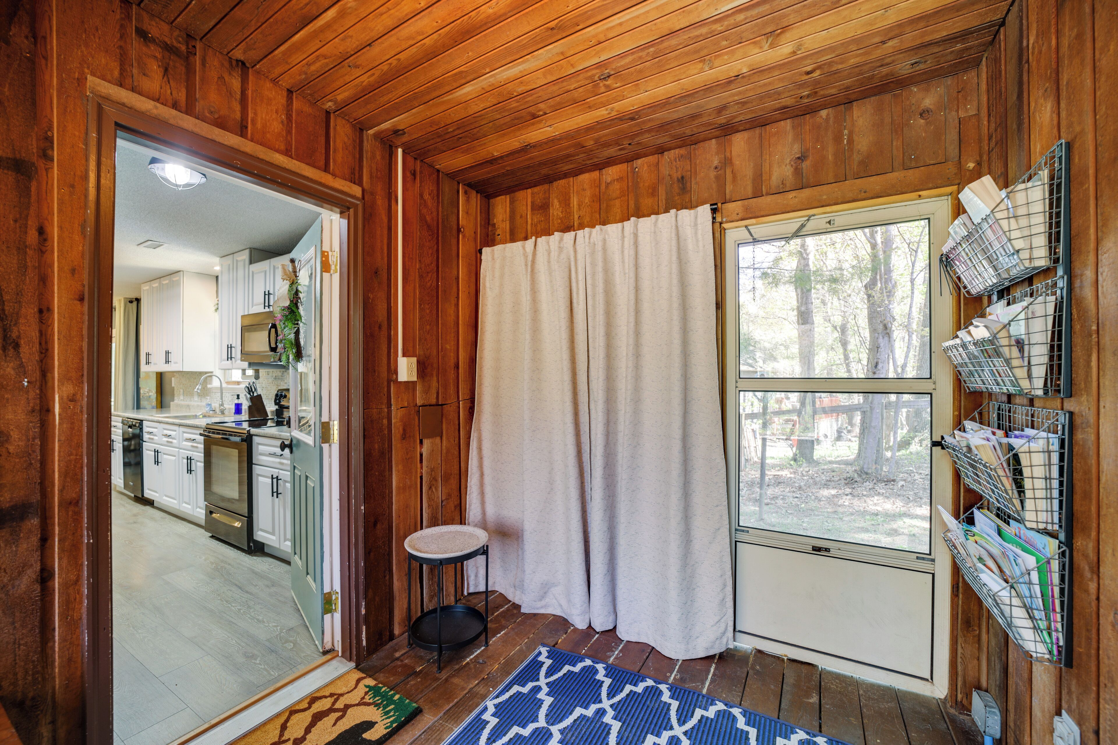 Un interior de madera con cocina, un taburete pequeño, una ventana con cortinas y una estantería con correo.