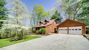 A two-story wooden house with a garage, surrounded by a well-maintained lawn and trees. - Peaceful Candler Cabin w/ Private Hot Tub! (Candler)