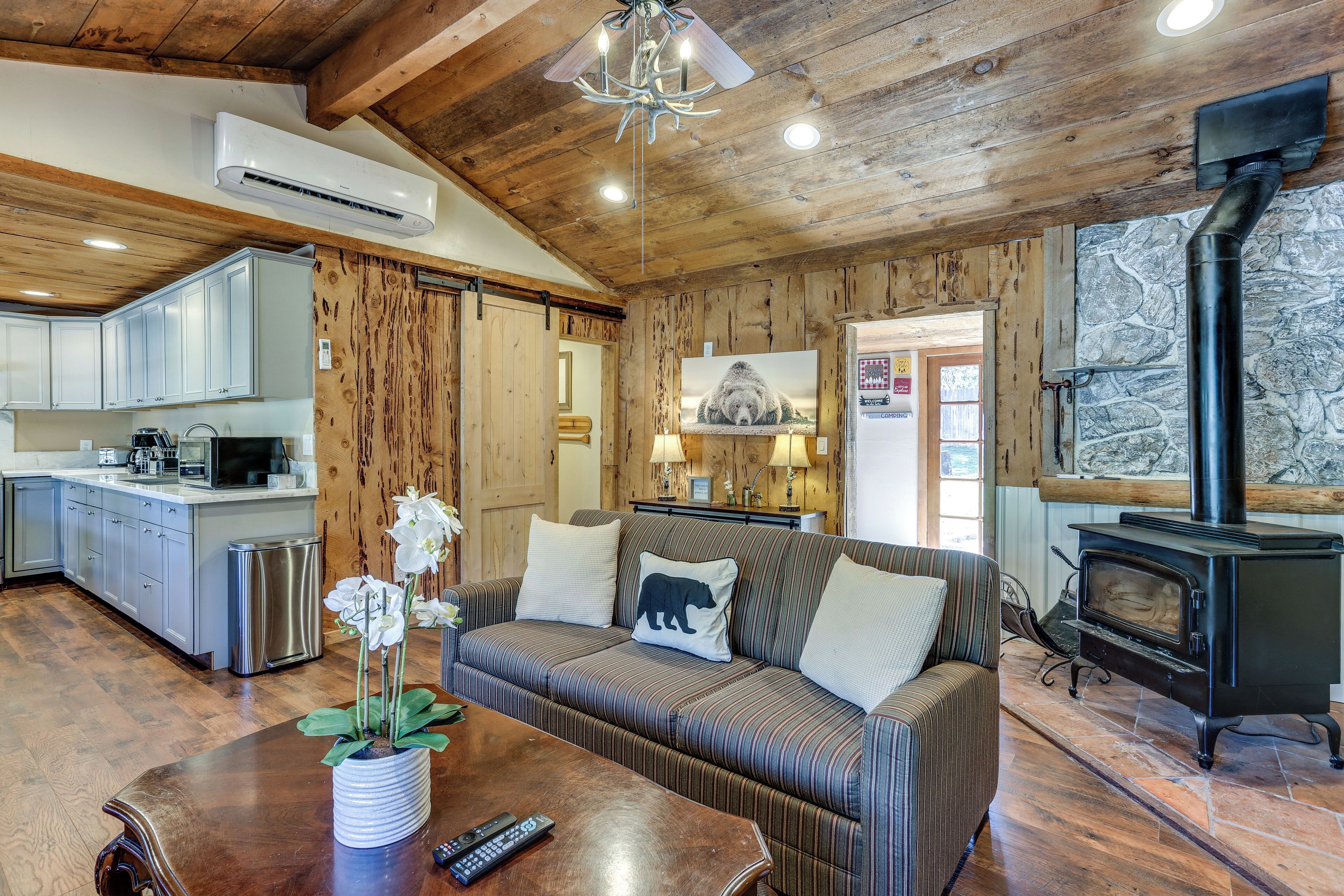 A cozy cabin interior with a wooden ceiling, a stone wall, a sofa with a bear pillow, a kitchen with white cabinets, and a wood-burning stove.