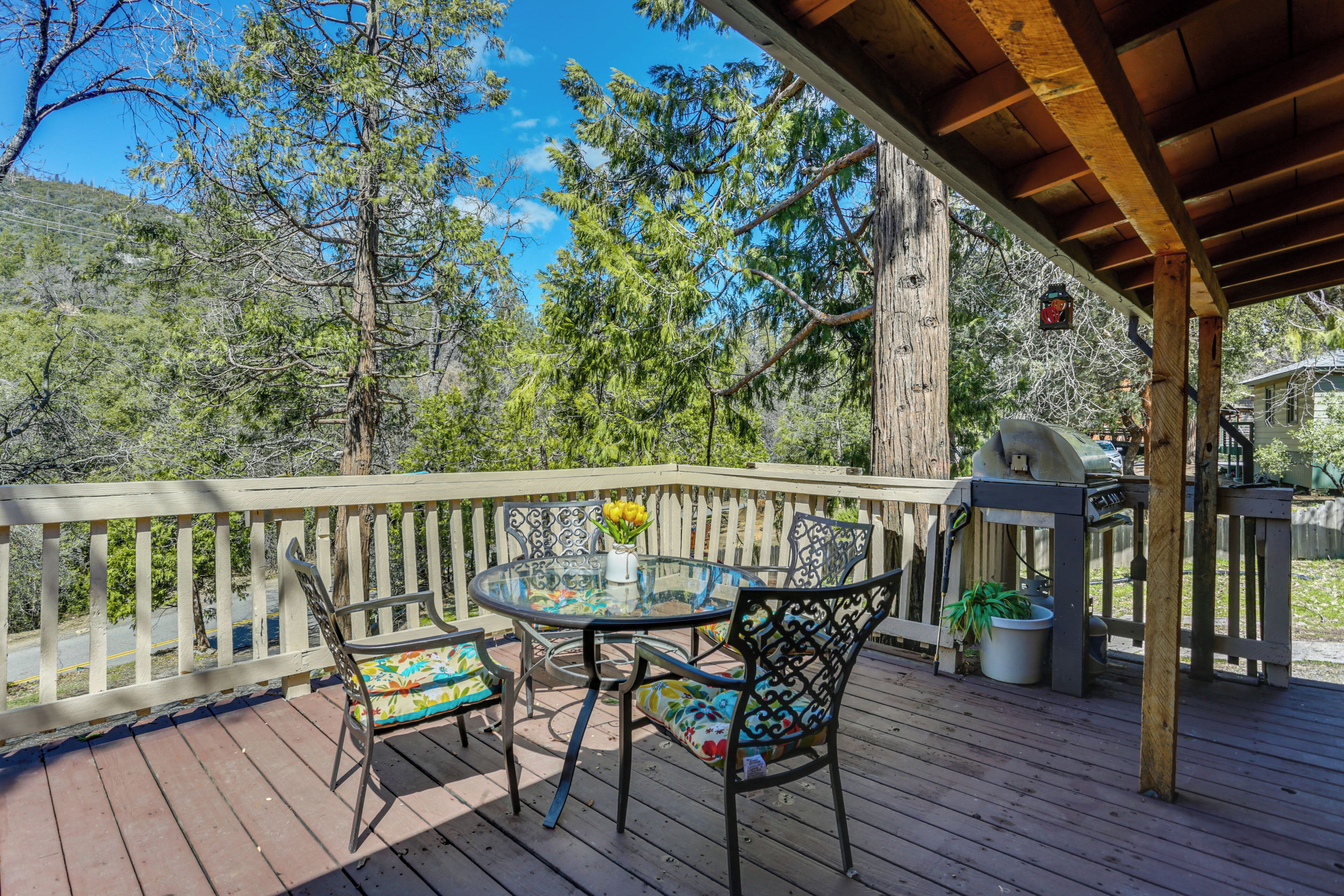 Une terrasse en bois avec une table et des chaises en verre, entourée d’une clôture blanche en bois, donnant sur une zone boisée.