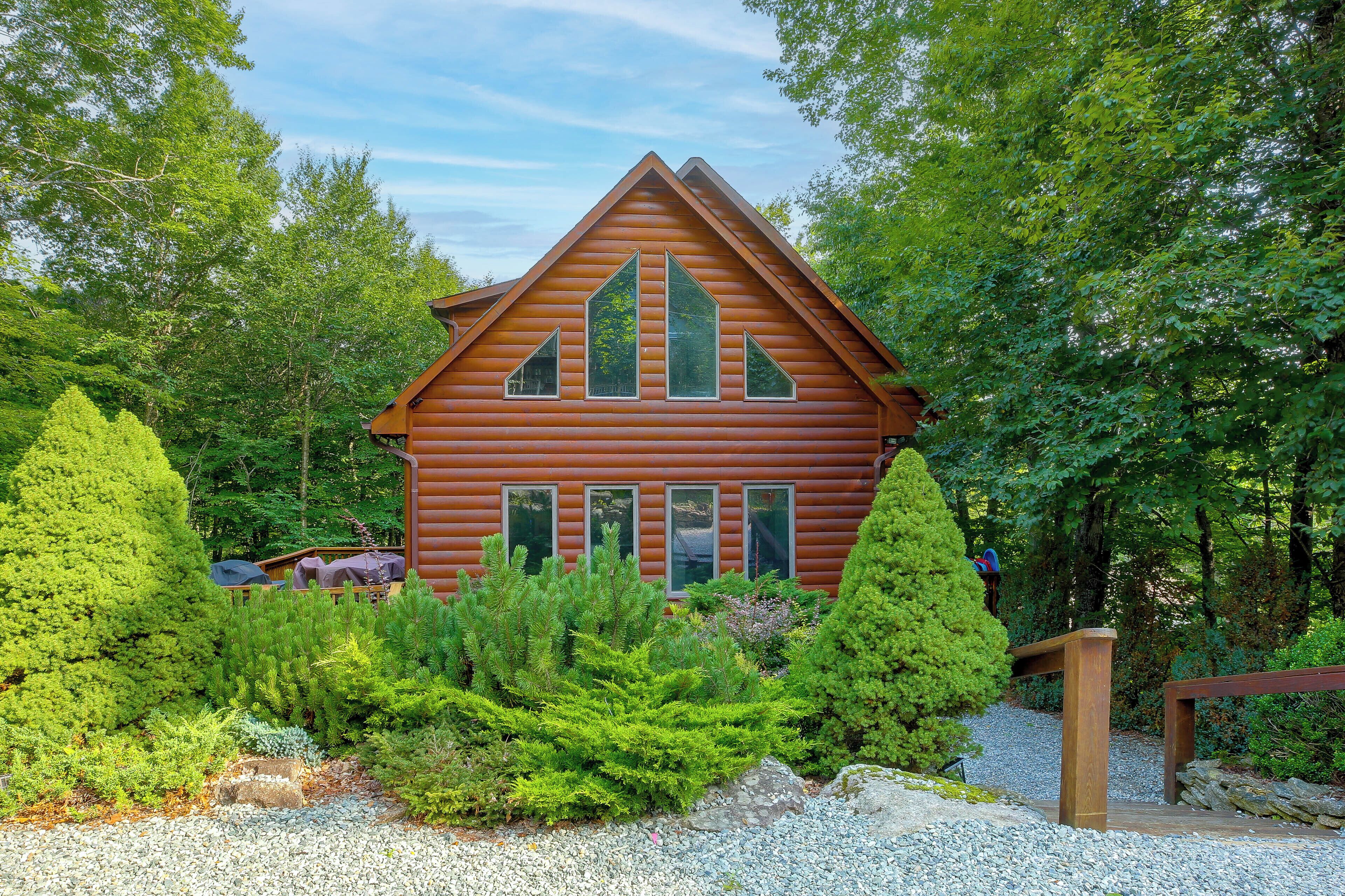 A log cabin with a gabled roof, surrounded by dense greenery and a gravel path.