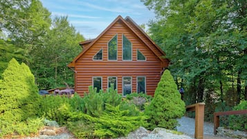 A log cabin with a gabled roof, surrounded by dense greenery and a gravel path.