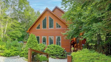 A log cabin with a steeply pitched roof, multiple windows, and surrounded by dense greenery.