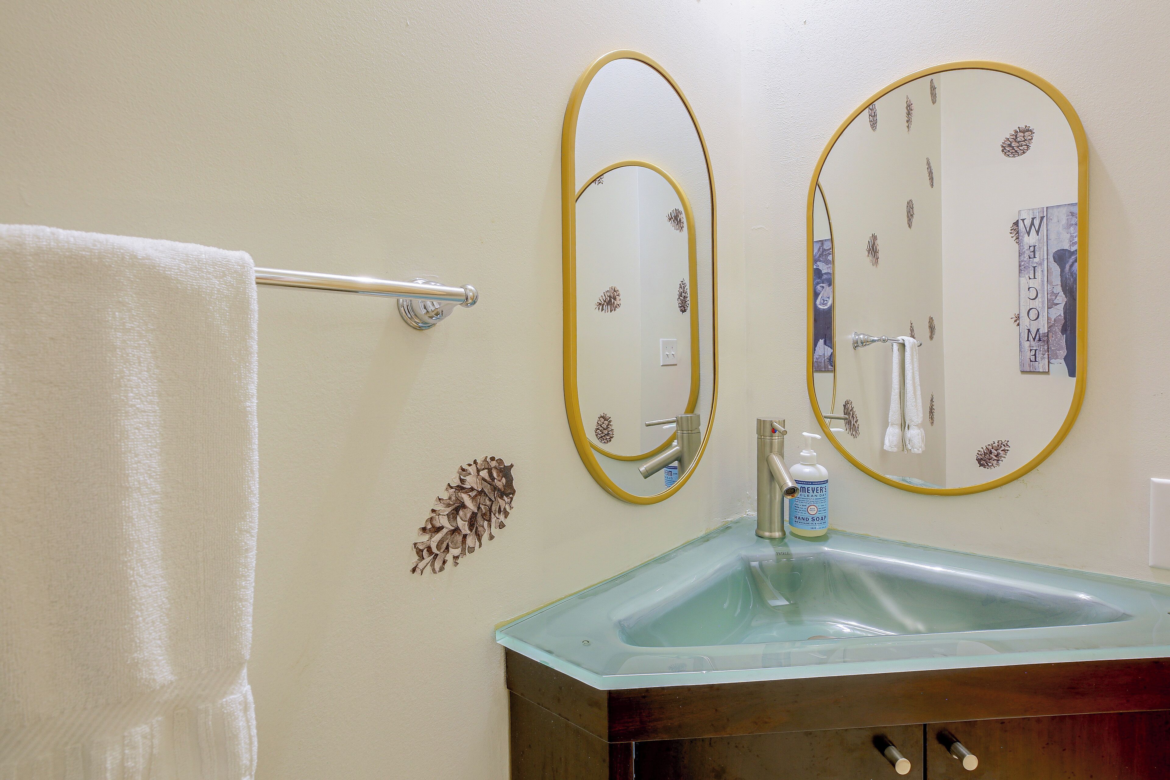 Ein Badezimmer mit einem Waschbecken aus Glas, zwei an der Wand befestigten Spiegeln mit goldfarbenen Rahmen, einem Handtuchhalter und einem 'WELCOME'-Schild.