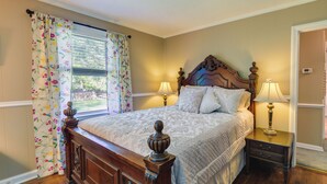A bedroom with a wooden bed, two bedside tables with lamps, a window with floral curtains, and a view of the outdoors. - Charming New Bern Cottage w/ Grill & Fire Pit! (New Bern)