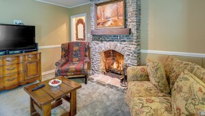 A living room with a stone fireplace, a television, a patterned armchair, and a sofa. - Charming New Bern Cottage w/ Grill & Fire Pit! (New Bern)