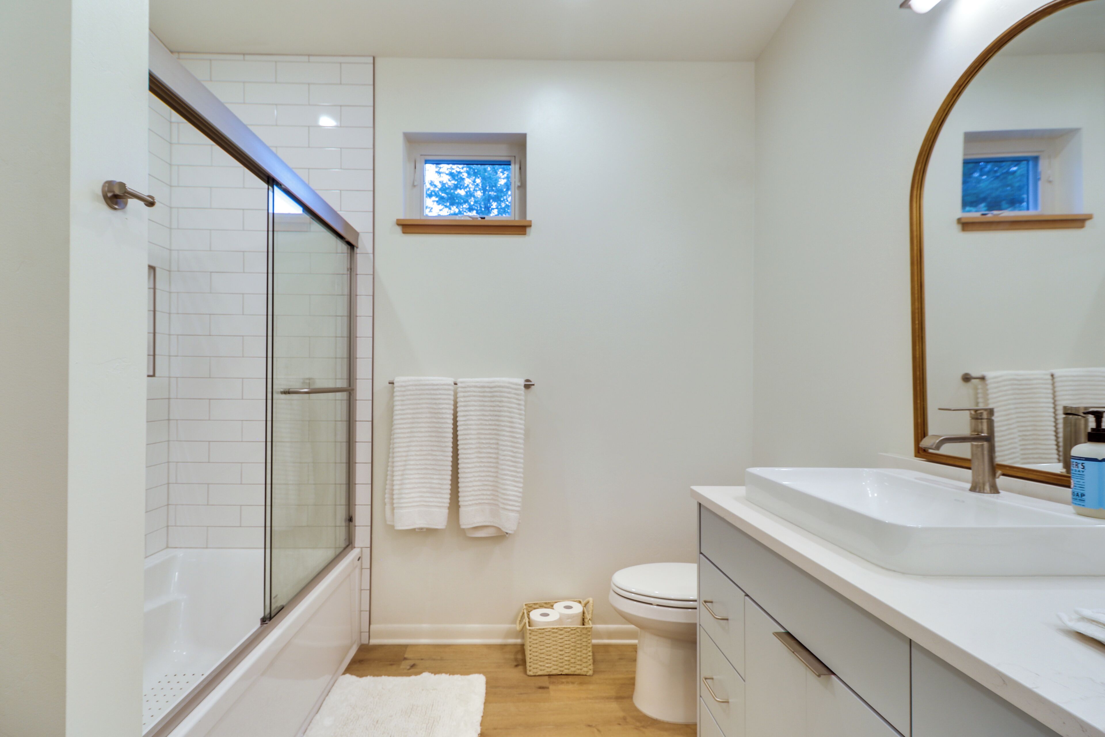 Un baño con ducha envuelta en vidrio, un lavamanos blanco, un inodoro y un ventanal pequeño.