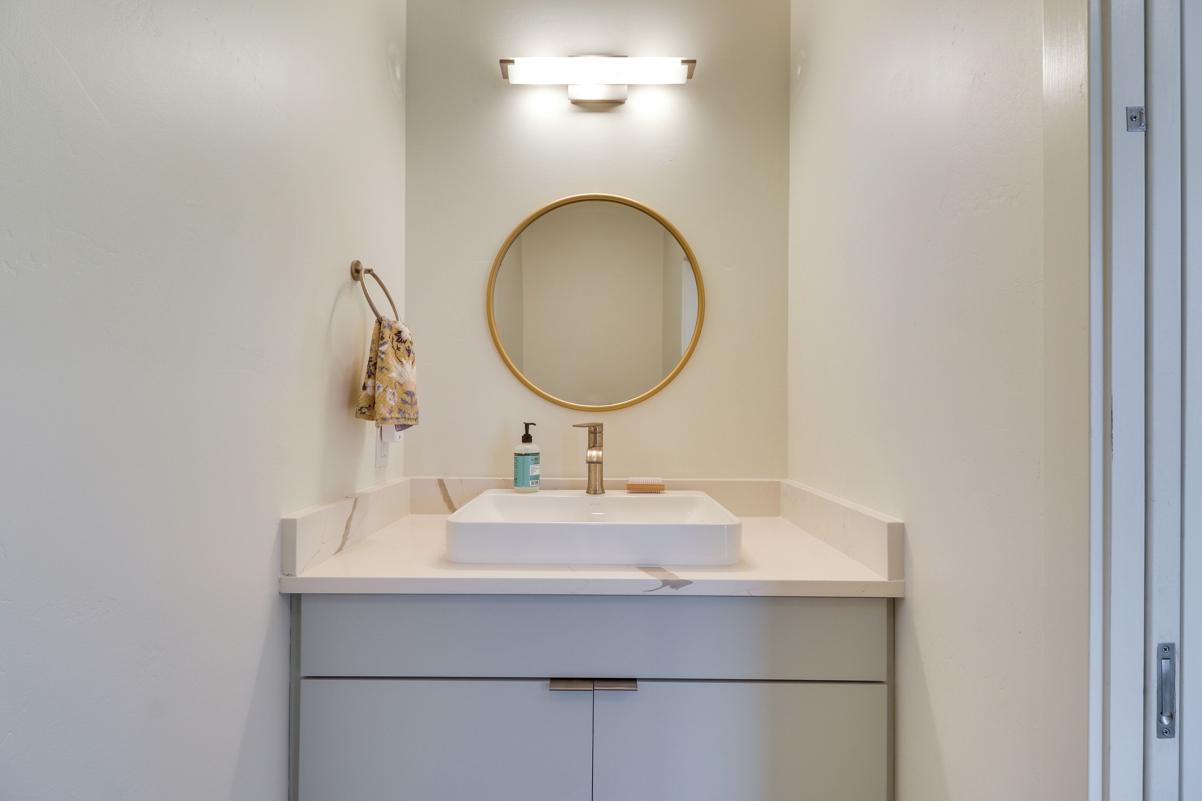A bathroom with a white sink, a round mirror, a handheld soap dispenser, and a small towel hanging on the wall.