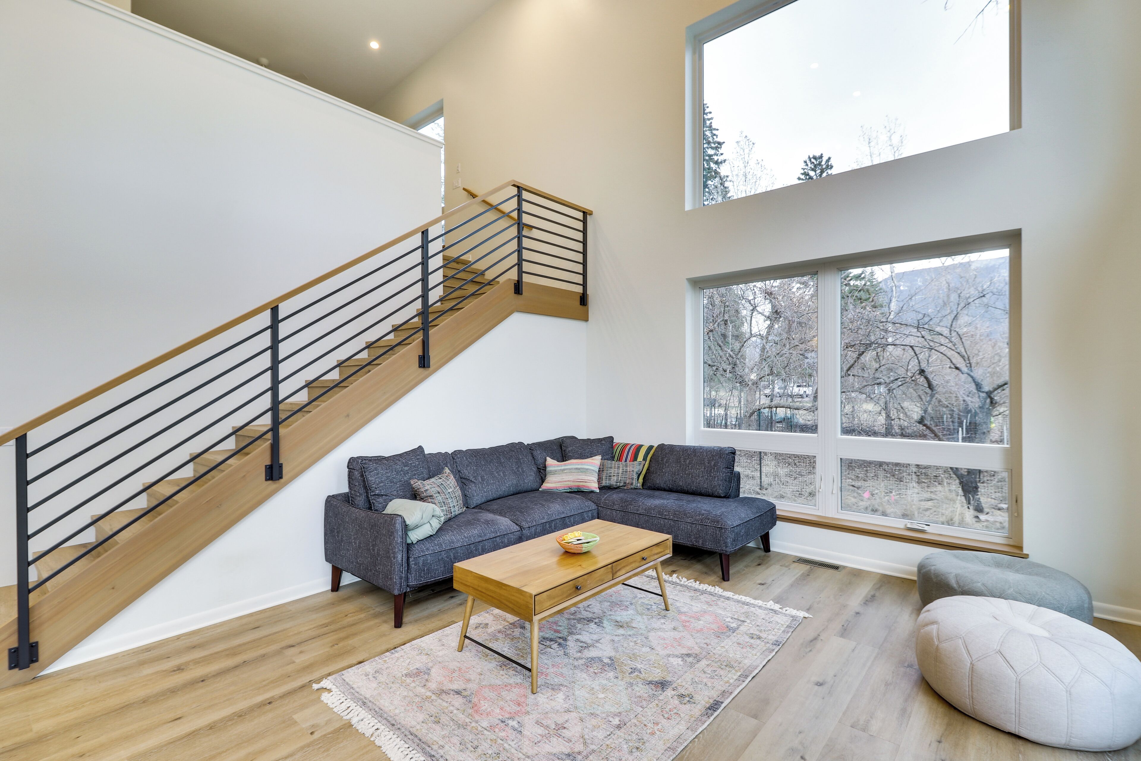 A modern living room with a wooden staircase, a sectional sofa, a coffee table, and a large window offering a view of trees.