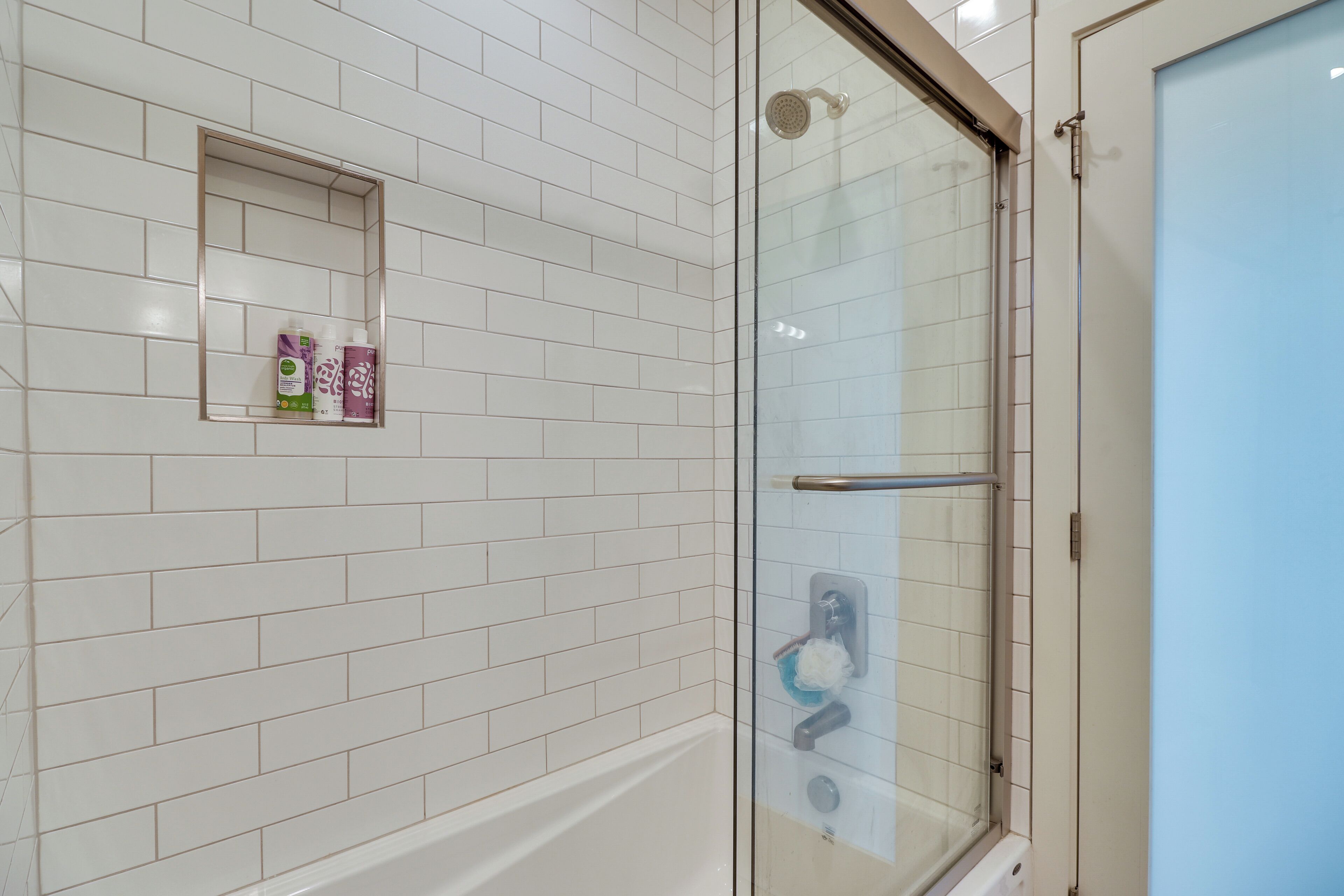 Un bagno con pareti piastrellate di bianco, un box doccia in vetro e un piccolo ripiano con articoli da toilette.