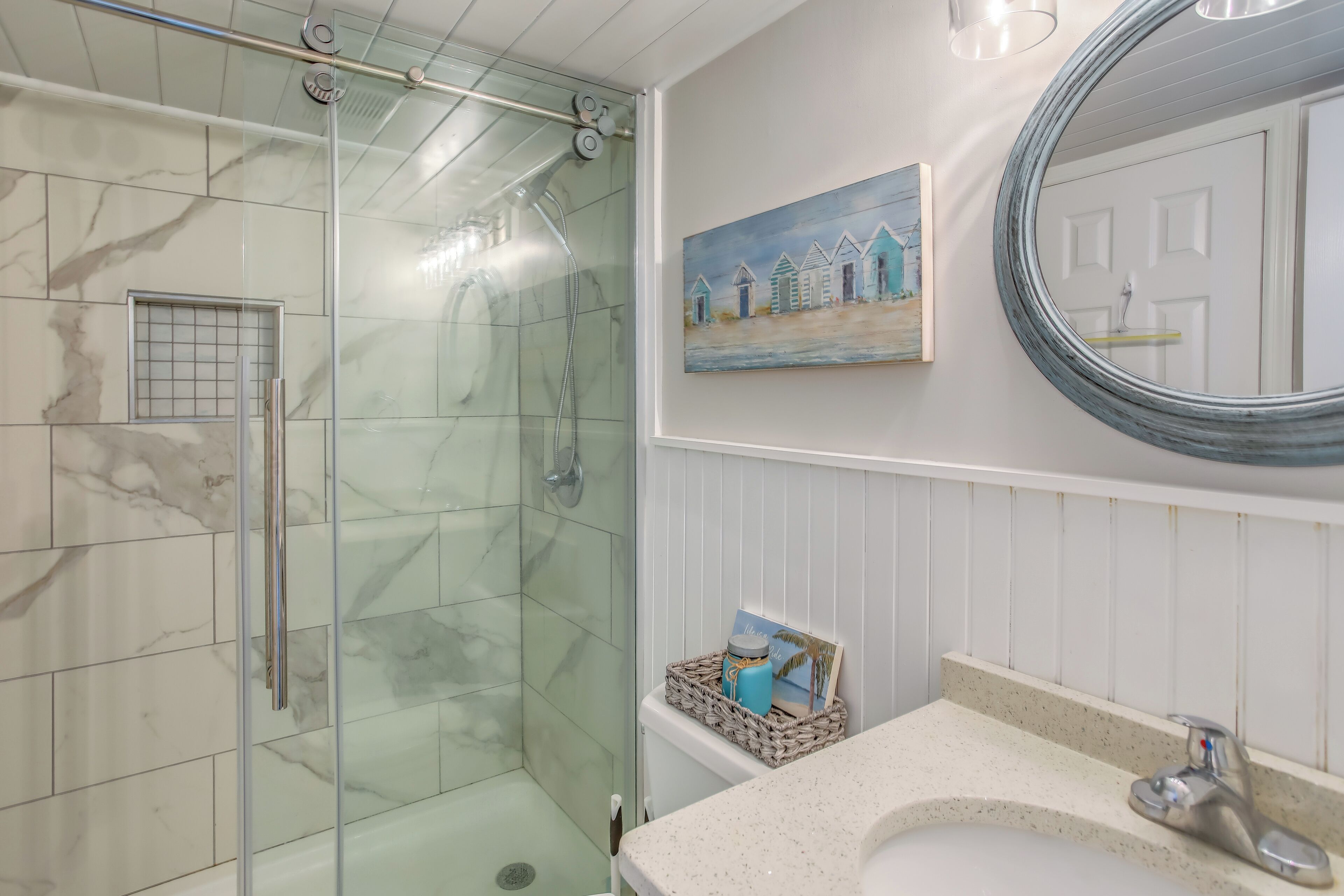 Un baño con ducha de vidrio, lavamanos blanco y un cuadro de casas de playa en la pared.