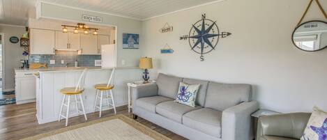Un salon chaleureux avec un canapé gris, une petite table blanche et un coin cuisine agrémenté d’une enseigne indiquant « MAISON DE PLAGE ».