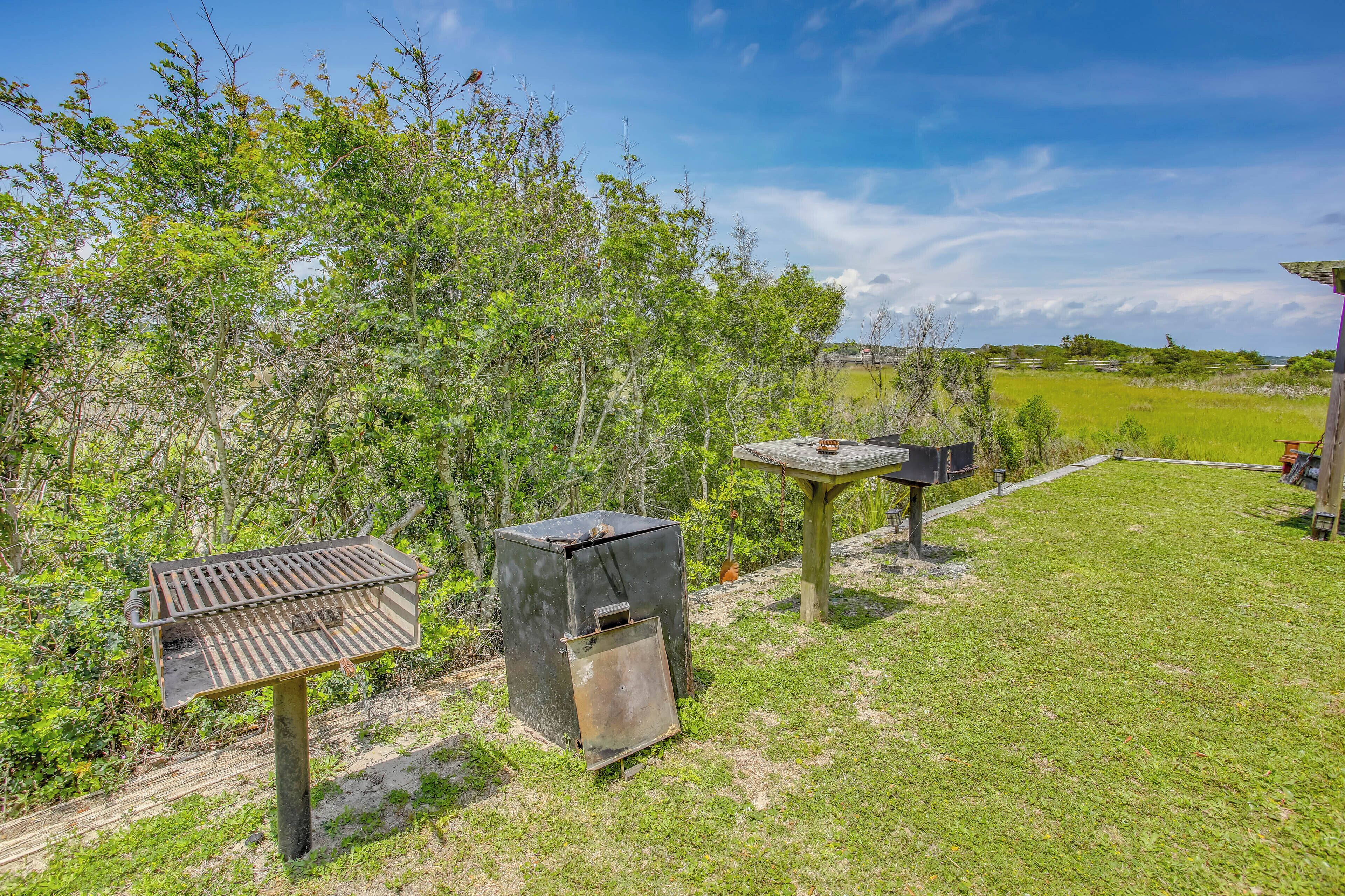 A barbecue grill, a trash bin, and a wooden table in a grassy outdoor area with trees in the background.