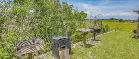 A barbecue grill, a trash bin, and a wooden table in a grassy outdoor area with trees in the background.