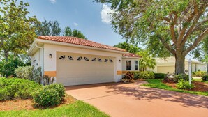 A single-story house with a red-tiled roof, a white garage door, and a well-maintained front yard with various shrubs and a large tree. - North Fort Myers Getaway w/ Resort Amenities! (North Fort Myers)