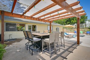 Um pátio externo com pérgola de madeira, uma mesa de jantar com cadeiras e uma área de piscina com espreguiçadeiras.