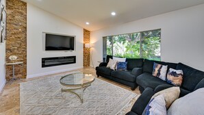A modern living room with a sectional sofa, a flat-screen TV mounted on a brick wall, a glass coffee table, and a large window with a view of greenery. - California Getaway w/ Private Outdoor Pool (Escondido)
