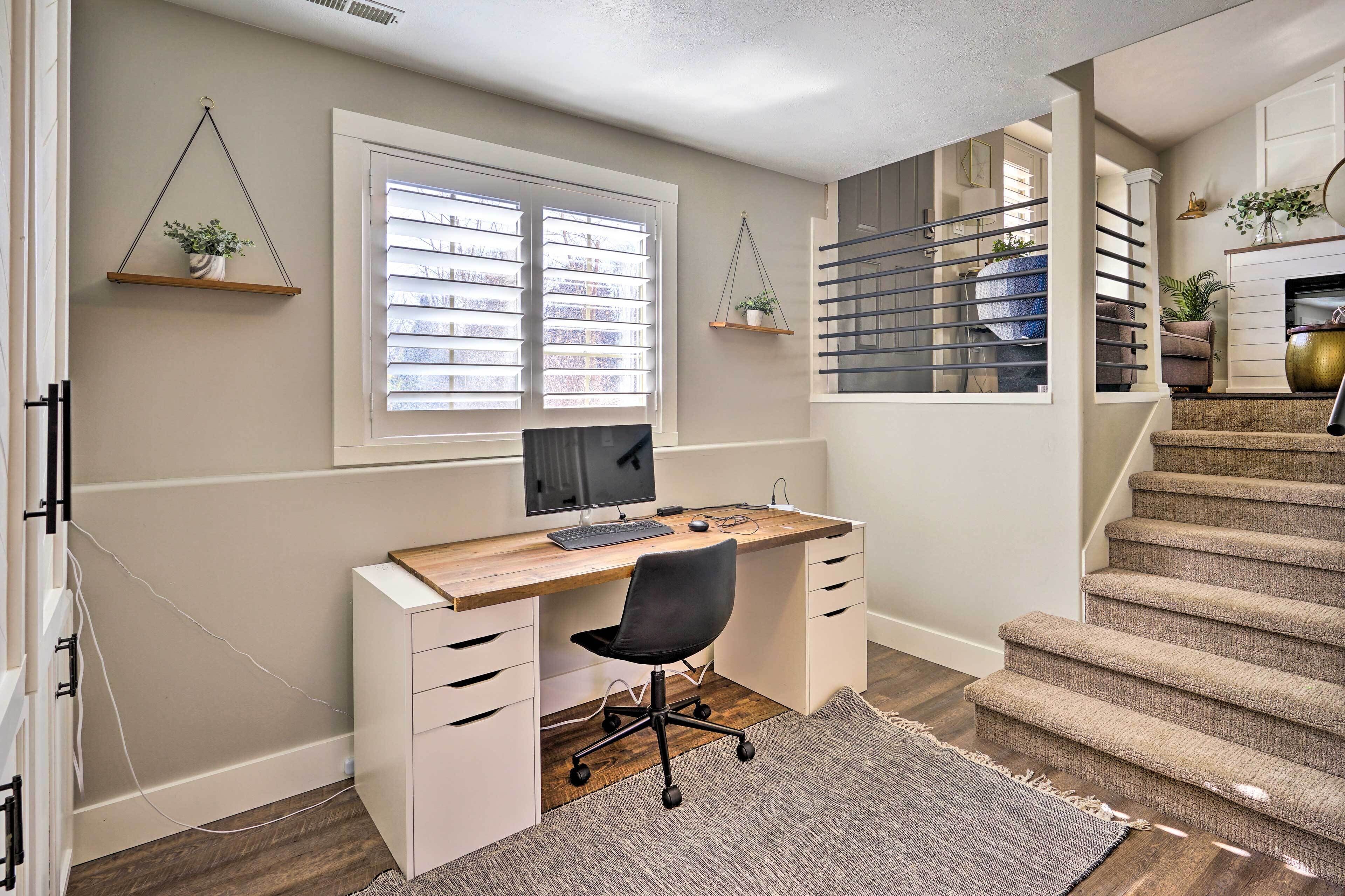 A home office with a desk, computer, and chair. There is a window with blinds, a staircase, and a rug on the floor.
