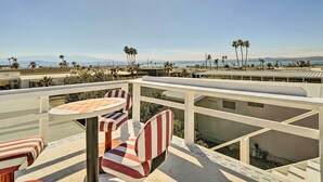 A balcony with striped chairs and a table, overlooking a beach with palm trees and mountains in the distance.