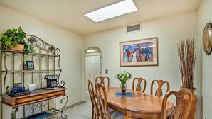A dining area with a wooden table, chairs, a metal coffee station, and a wall-mounted shelf with plants.