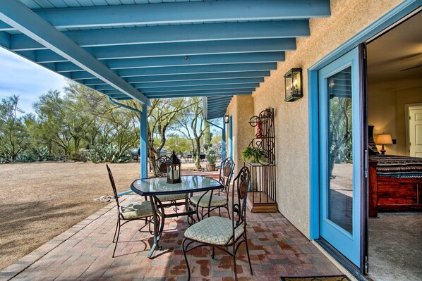 Un patio cubierto con una mesa y sillas, una puerta que da al interior, y vistas del paisaje circundante.