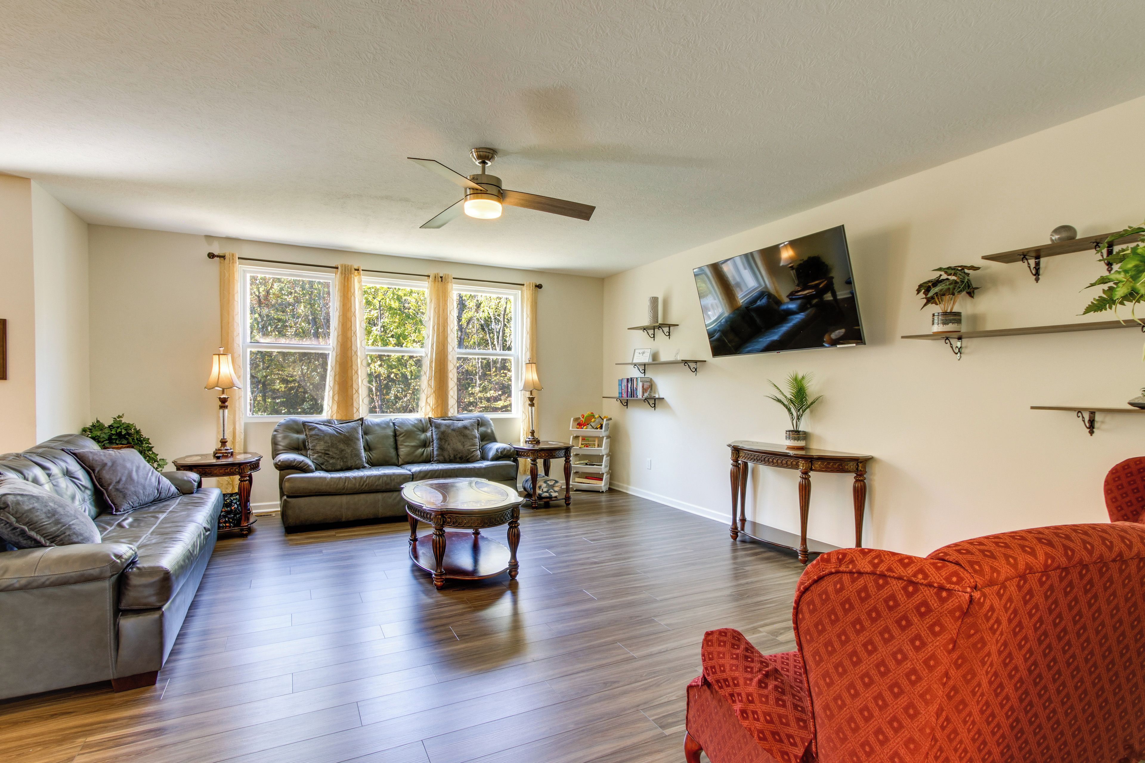 A spacious living room with hardwood flooring, a ceiling fan, a large window with curtains, a flat-screen TV mounted on the wall, and several pieces of furniture including sofas and a coffee table.