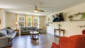 A spacious living room with hardwood flooring, a ceiling fan, a large window with curtains, a flat-screen TV mounted on the wall, and several pieces of furniture including sofas and a coffee table.