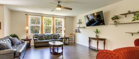 A spacious living room with hardwood flooring, a ceiling fan, a large window with curtains, a flat-screen TV mounted on the wall, and several pieces of furniture including sofas and a coffee table.