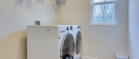Een wasserette met een voorlader wasmachine en droger, een raam, een plank met schoonmaakmiddelen, en een prullenbak.