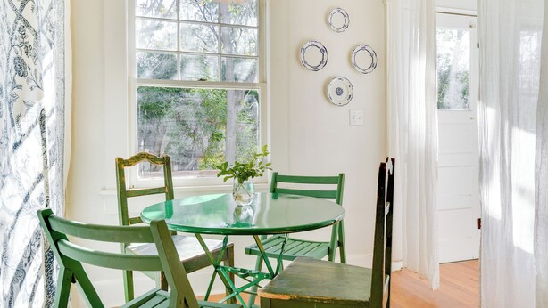 A dining area with a round glass table, green chairs, a vase with flowers, and a window with a view of trees.