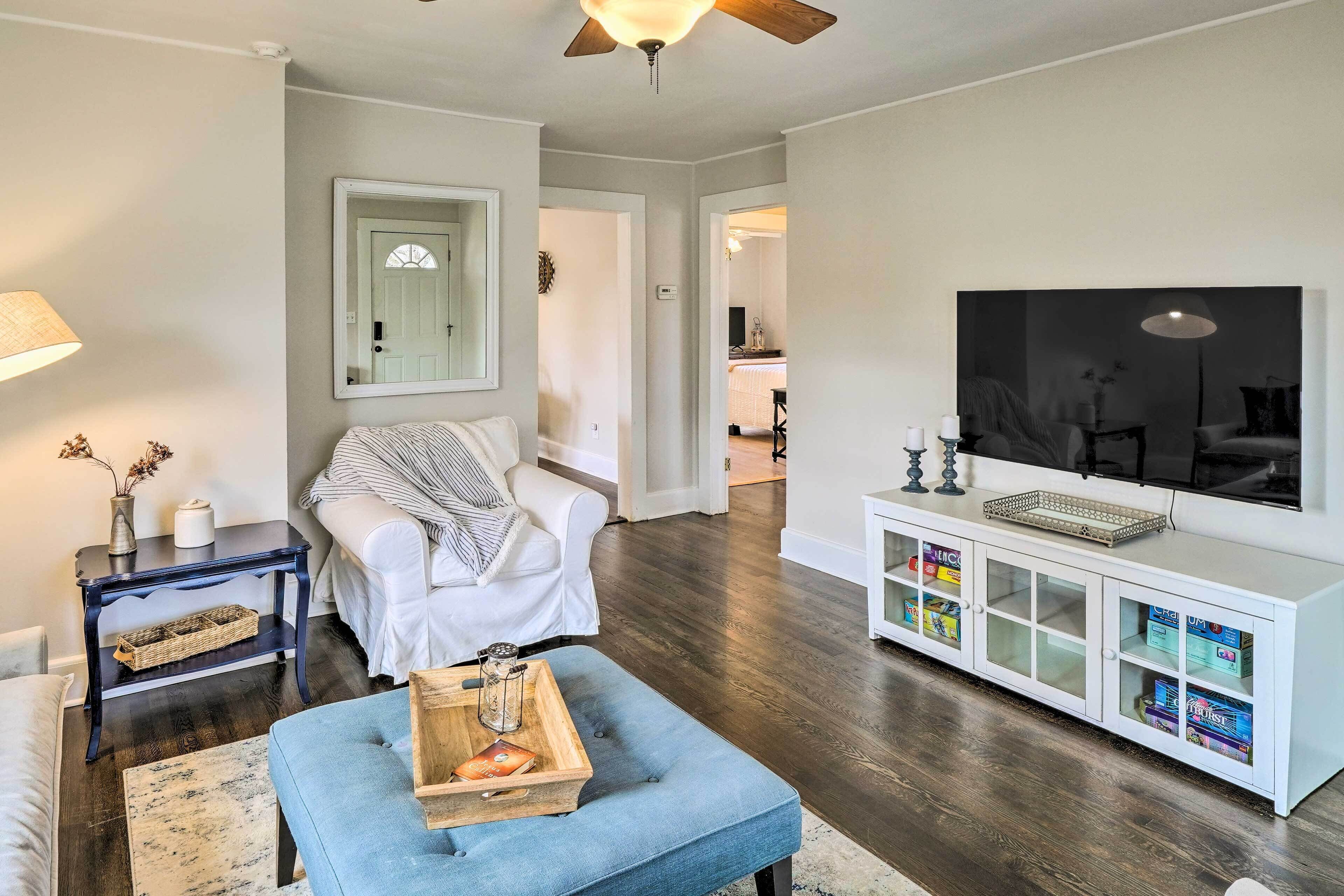 Un salon avec un canapé blanc, un pouf bleu, une table basse en bois foncé, un meuble de télévision blanc avec des portes en verre et un ventilateur de plafond.