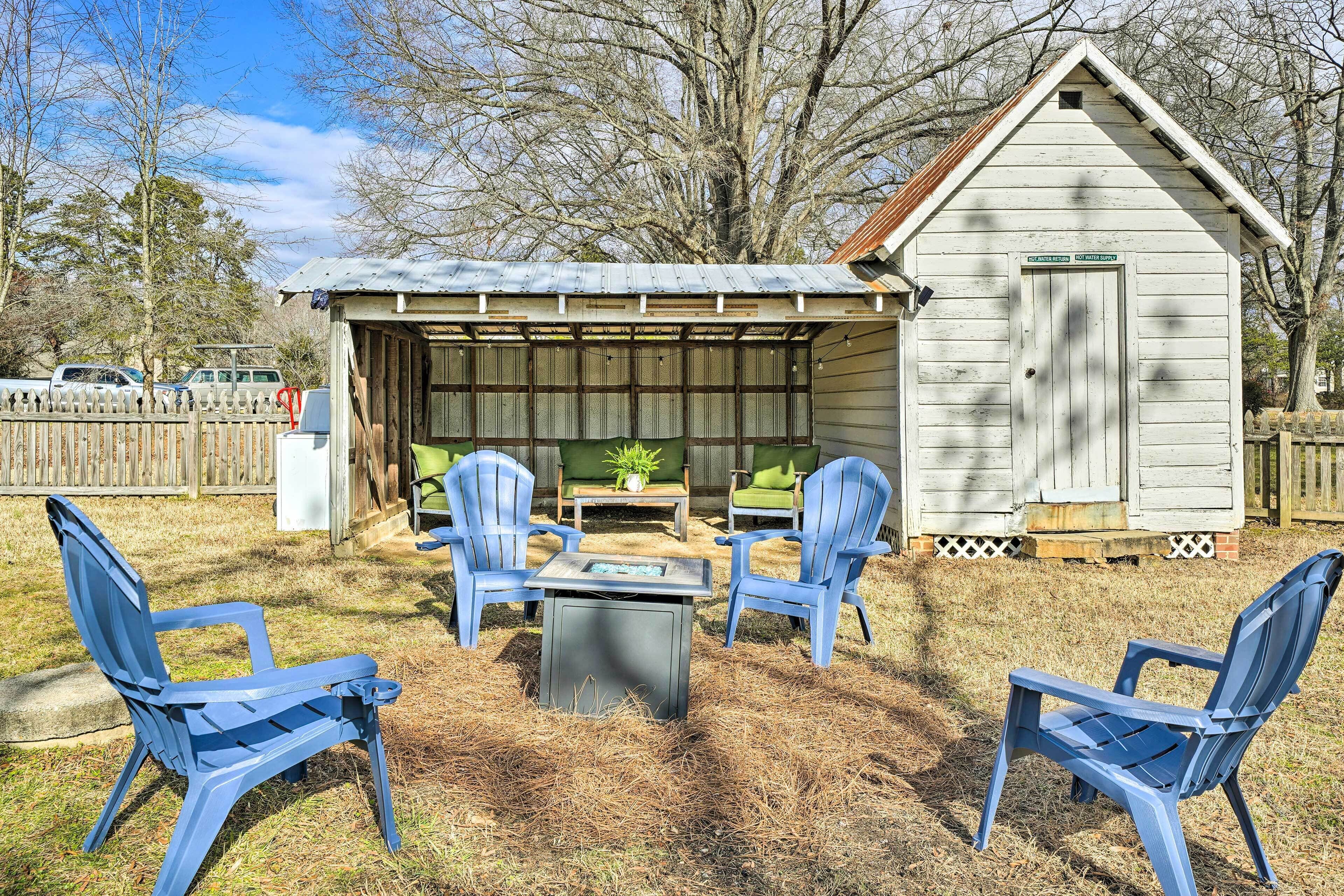 Un espacio de estar al aire libre cubierto con una mesa y cuatro sillas Adirondack azules, un cobertizo pequeño y una valla de madera al fondo.